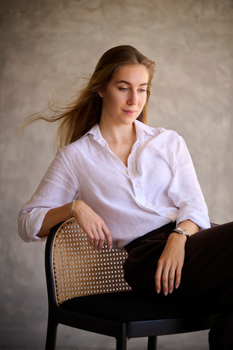 Professional portrait of a woman seated on a cane chair against a textured grey wall